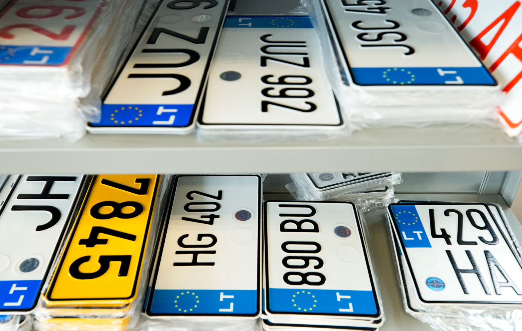 Lietuvos Europos Sąjungos numerių lentelių kolekcija, išdėstyta lentynose. Matomi įvairūs numeriai, tokie kaip JUZ 962, ZJH 7845 (geltoname fone), HG 402, 890 BU, JSC 459 ir LT 429 HA.