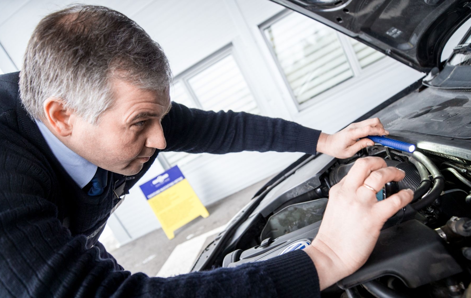 Asmuo apžiūri automobilio variklį pro atidarytą kapotą, naudodamas prožektorių; fone – pastatas su langais ir geltonas įspėjamasis ženklas.