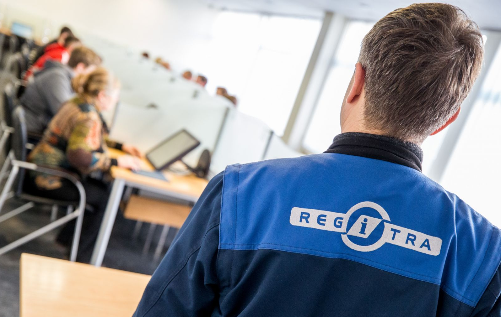 Asmuo su mėlyna striuke, kurios nugaroje matomas „REGITRA“ logotipas, fone – žmonės prie kompiuterių biuro ar egzamino aplinkoje.