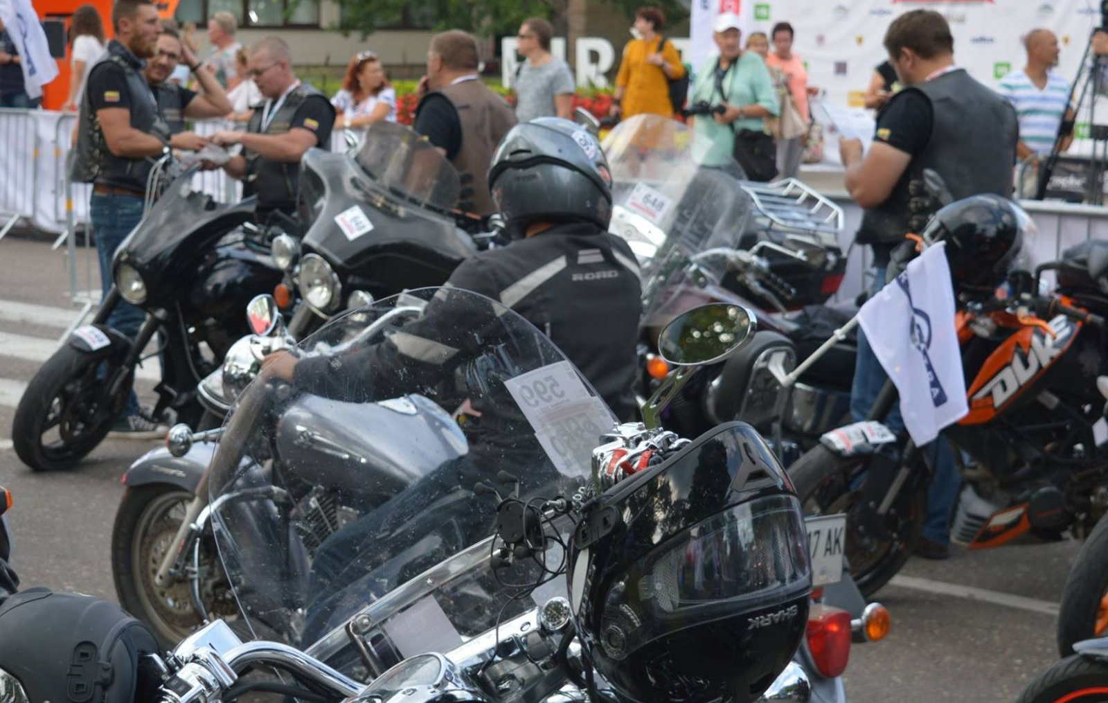 Grupė motociklininkų su šalmais ir apsaugomis stovi prie glaudžiai sustatytų motociklų, fone matomi žmonės ir bendravimas.