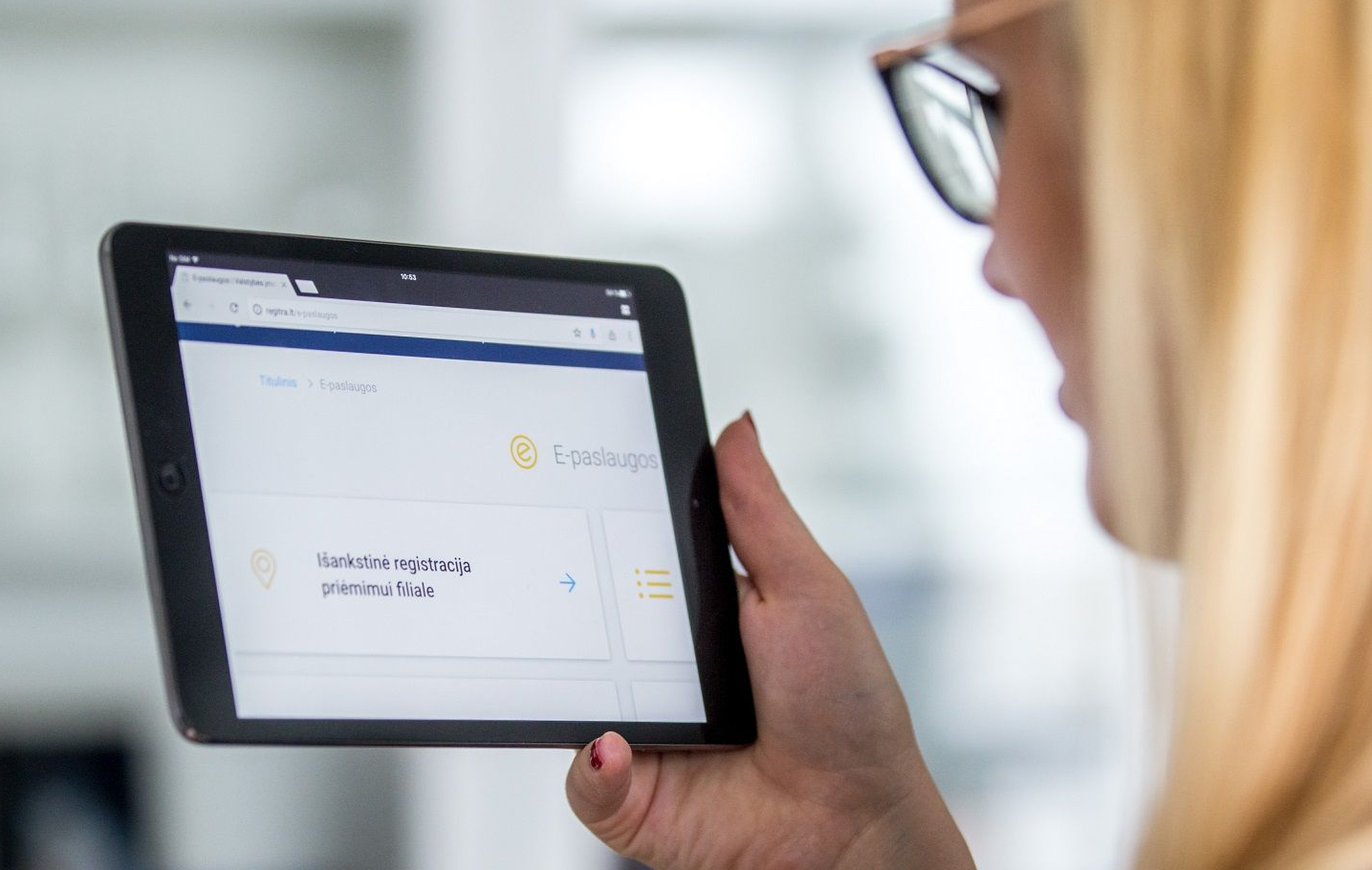 Asmuo laiko planšetę, kurios ekrane matomas puslapis su užrašais „E-paslaugos“ ir „Išankstinė registracija priėmimui filiale