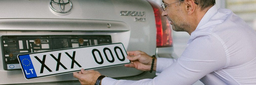 Asmuo su baltais marškiniais ir mėlynomis kelnėmis laiko ženklą „XXX 000“ prie sidabrinės „Toyota“ automobilio