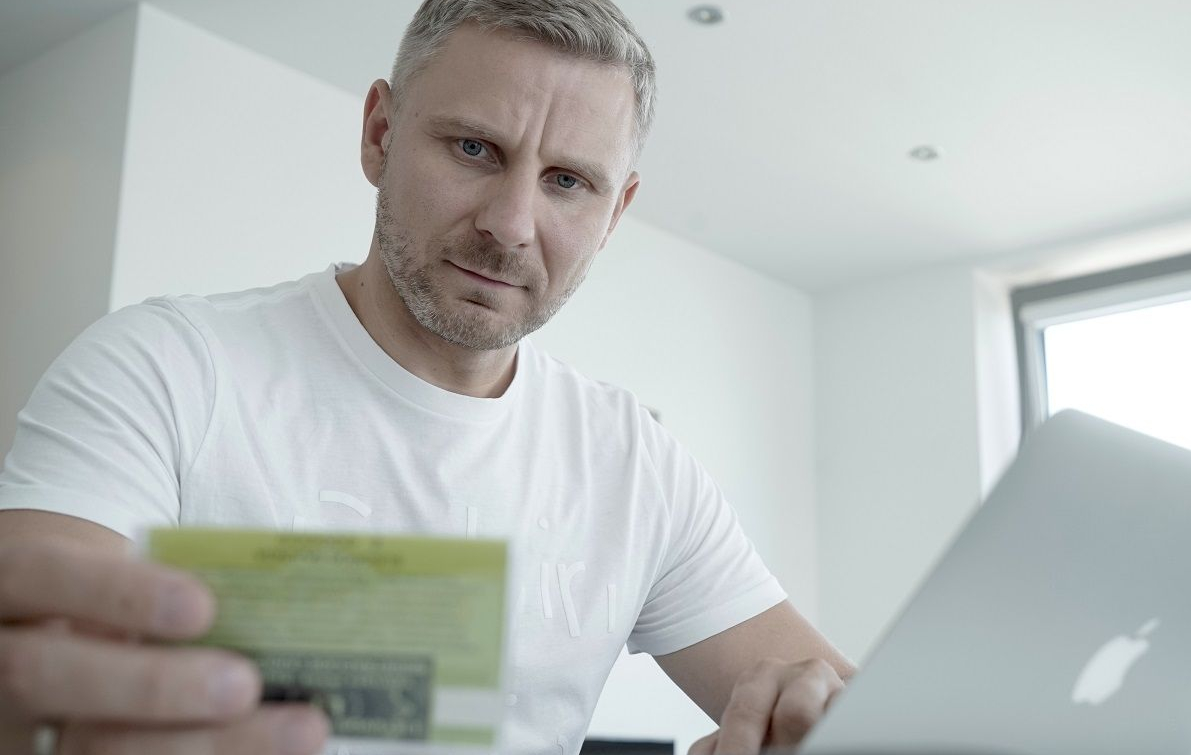 Vyras su baltais marškinėliais žiūri į žalią dokumentą, tikriausiai registracijos liudijimą.