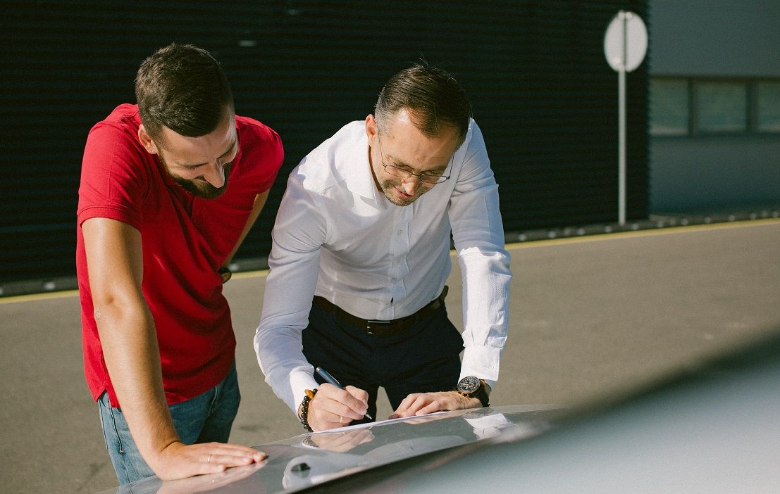 Du žmonės stovi prie automobilio lauke, vienas rašo ant popieriaus, padėto ant automobilio.