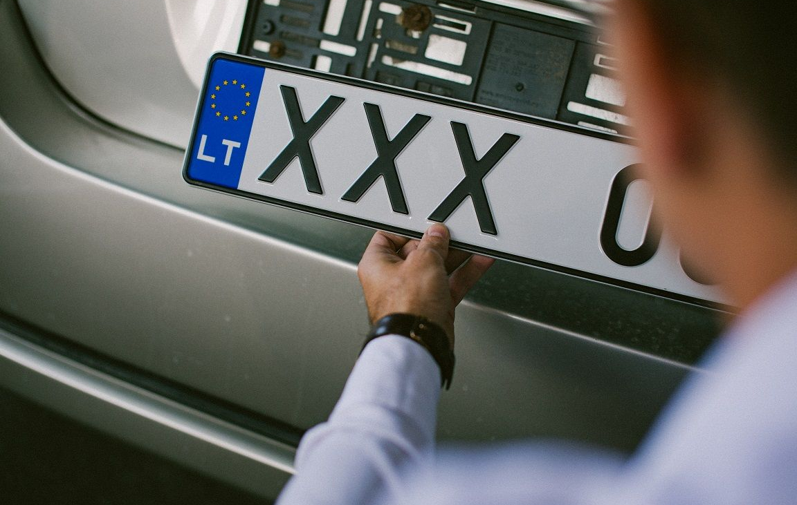 Asmuo su baltais marškiniais ir mėlynomis kelnėmis tvirtina arba nuima numerio lentelę nuo sidabrinio „Toyota“ automobilio. Numeris – XXX 000, su ES simboliu ir „LT“ žyma.
