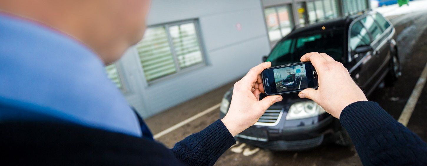 Nuotraukoje matomas žmogus, fotografuojantis juodą automobilį išmaniuoju telefonu. Automobilis stovi priešais pastatą su dideliais langais, o telefono ekrane matyti fotografuojamas automobilis.