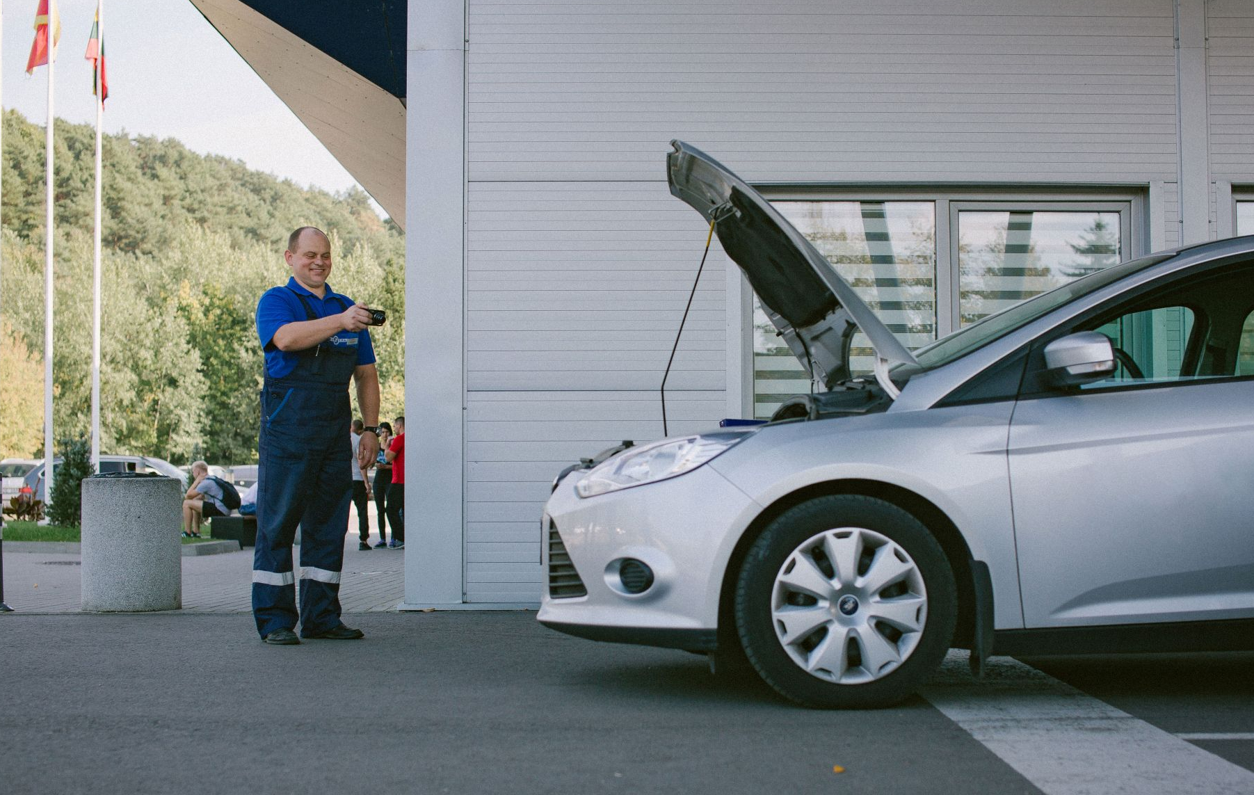 Asmuo stovi šalia sidabrinio automobilio su atidarytu kapotu ir rodo ar gestikuliuoja jo link. Scena vyksta lauke, fone – pastatas su dideliais langais, matomos vėliavos ir keli žmonės tolumoje