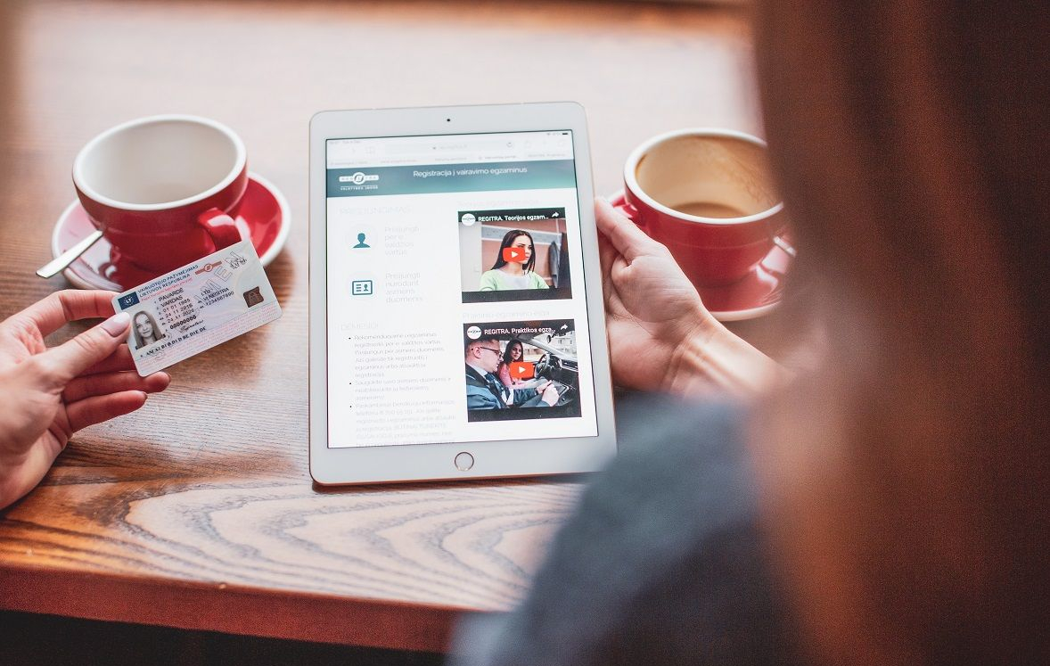 Asmuo laiko vairuotojo pažymėjimą vienoje rankoje, o planšetę – kitoje. Planšetės ekrane matomas tinklalapis su tekstu ir vaizdais. Ant stalo – du raudoni puodeliai, vieną jų laiko kitas asmuo