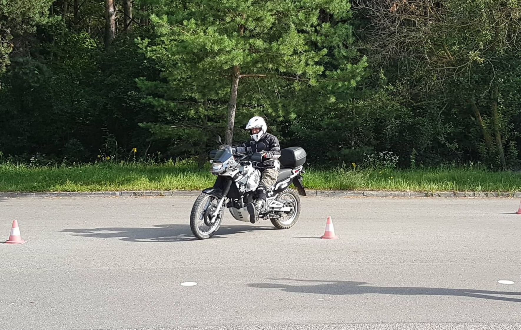 Asmuo su apsaugine motociklininko apranga važiuoja motociklu grįstu paviršiumi šalia mažo kelio kūgio. Fone – žali medžiai ir žolė.