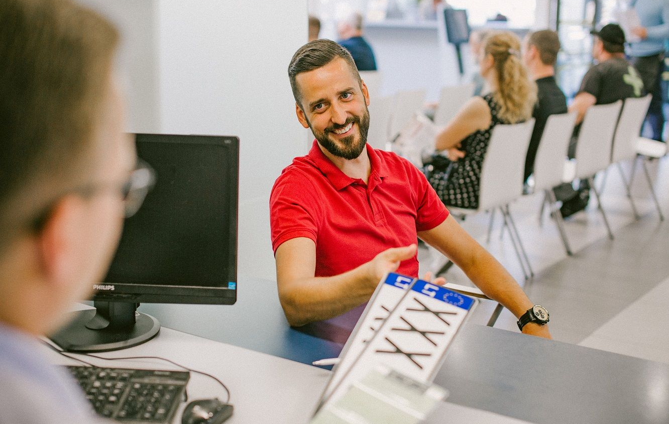 Vyras su raudonais polo marškinėliais sėdi prie stalo ir gauna transporto priemonės numerių lenteles. Ant stalo – kompiuterio monitorius, fone matomi kiti žmonės.