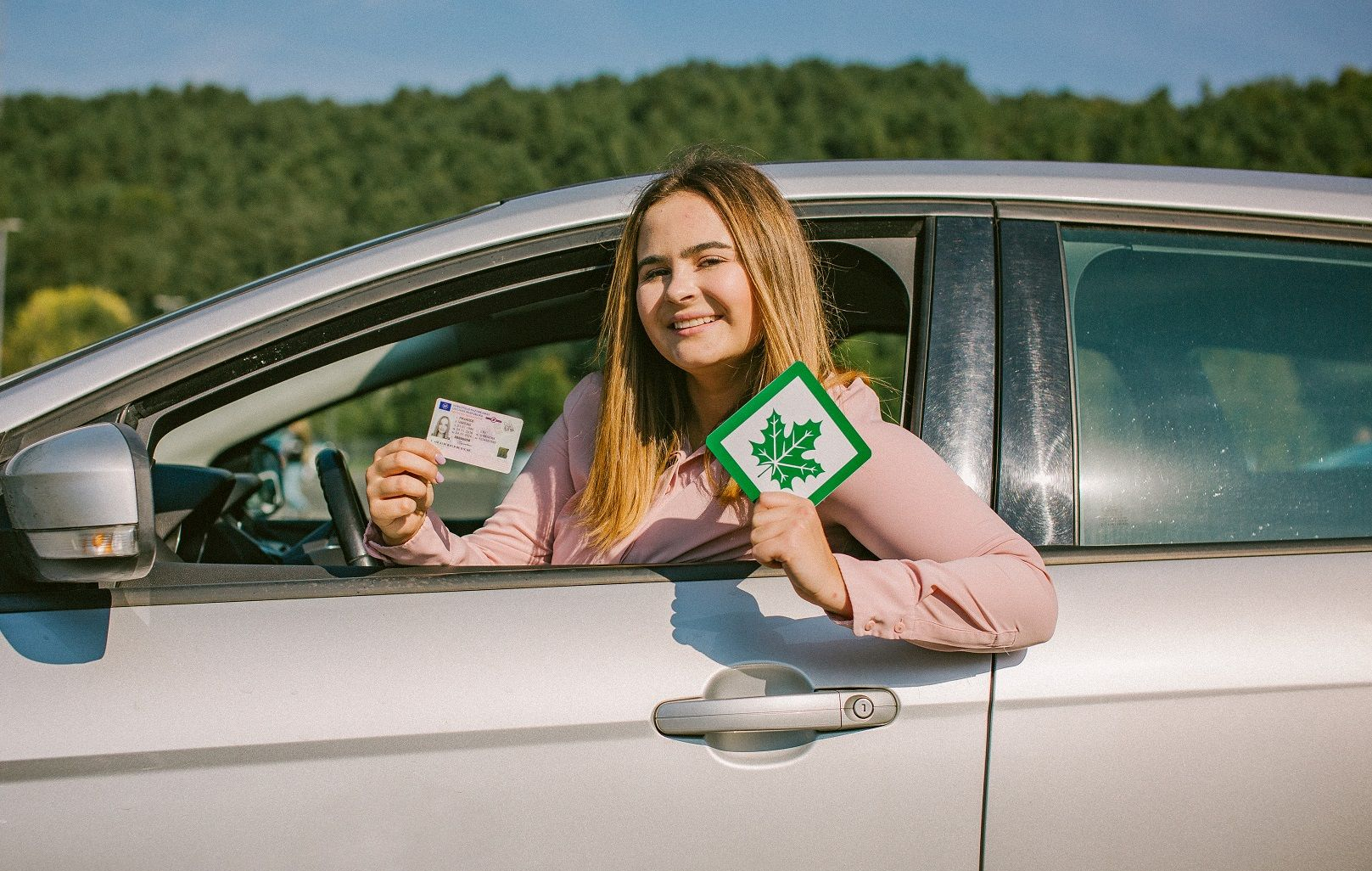 Asmuo sėdi sidabrinio automobilio vairuotojo vietoje, vienoje rankoje laiko vairuotojo pažymėjimą, kitoje – žalią klevo lapo ženklą. Fone matoma žaluma ir medžiai