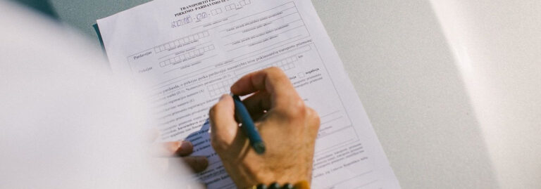 Asmuo rašo rašikliu ant dokumento su keliais laukeliais ir žymimais langeliais.