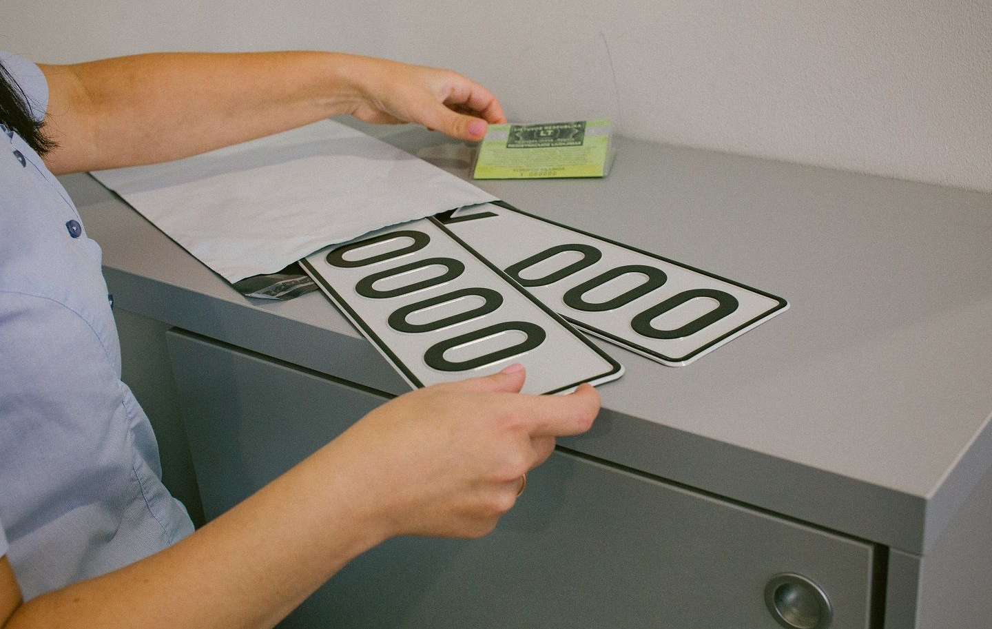 Asmuo stovi prie pilko paviršiaus ir laiko dvi stačiakampes lenteles su numerio ženklaus „0000“ bei registracijos liudijimą