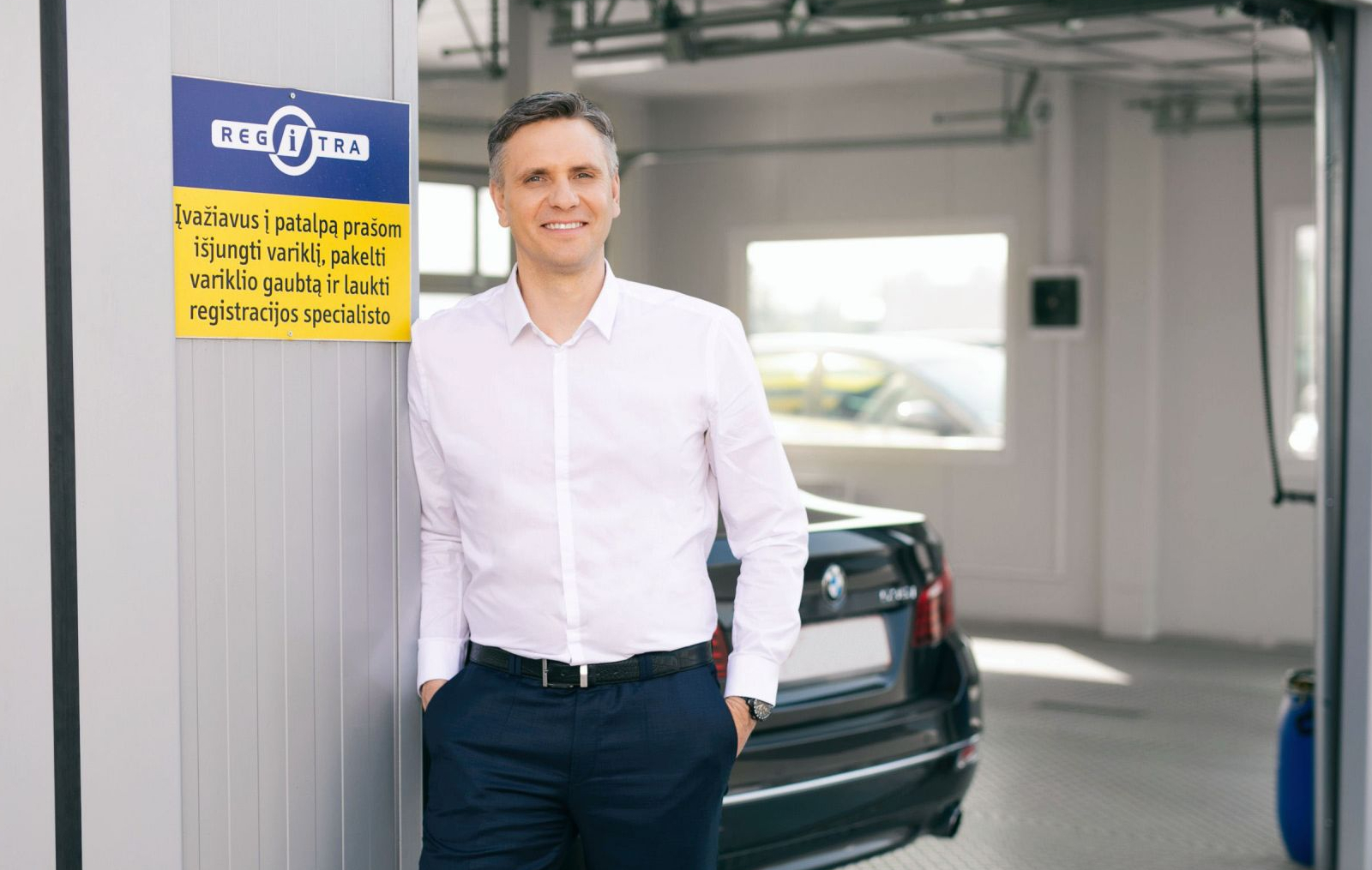 Asmuo su baltais marškiniais ir tamsiomis kelnėmis stovi garaže ar transporto registracijos patalpoje prie juodo automobilio. Šalia matomas ženklas su užrašu: „REGITRA. Įvažiavus į patalpą prašom išjungti variklį, pakeisti variklio gaubtą ir laukti registracijos specialisto.“