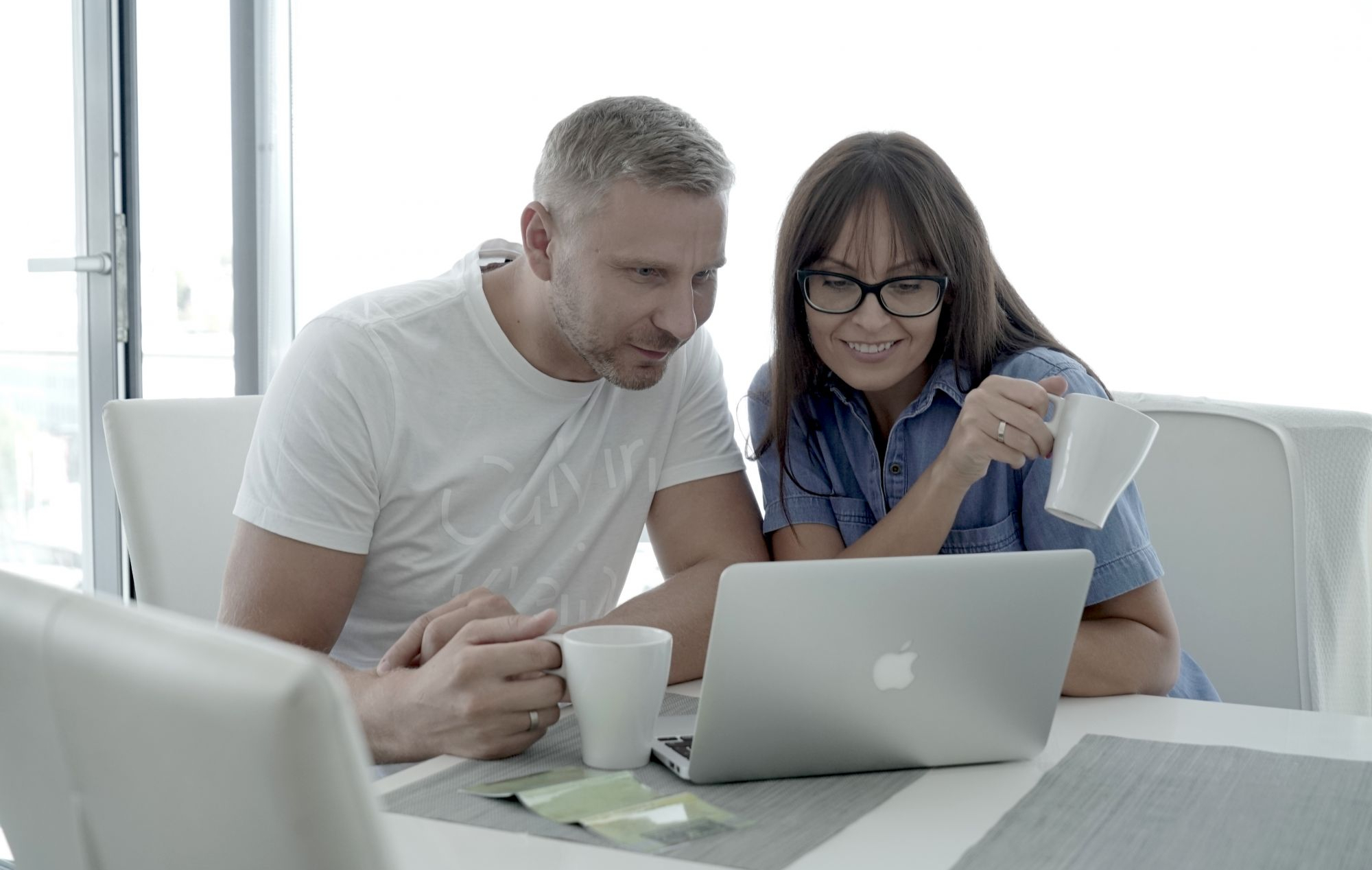 Du asmenys sėdi prie stalo ir žiūri į „Apple“ nešiojamąjį kompiuterį. Vienas laiko baltą puodelį, ant stalo yra dar du puodeliai ir keli paketėliai.