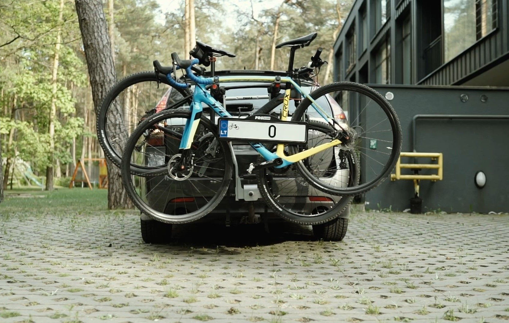 Automobilio galas su dviem dviračiais pritvirtintais prie laikiklio. Transporto priemonė stovi ant grįsto paviršiaus su žole tarp akmenų. Matoma numerio lentelė su „0“ ir „LT“ žyma. Fone – medžiai ir pastato dalis dešinėje.