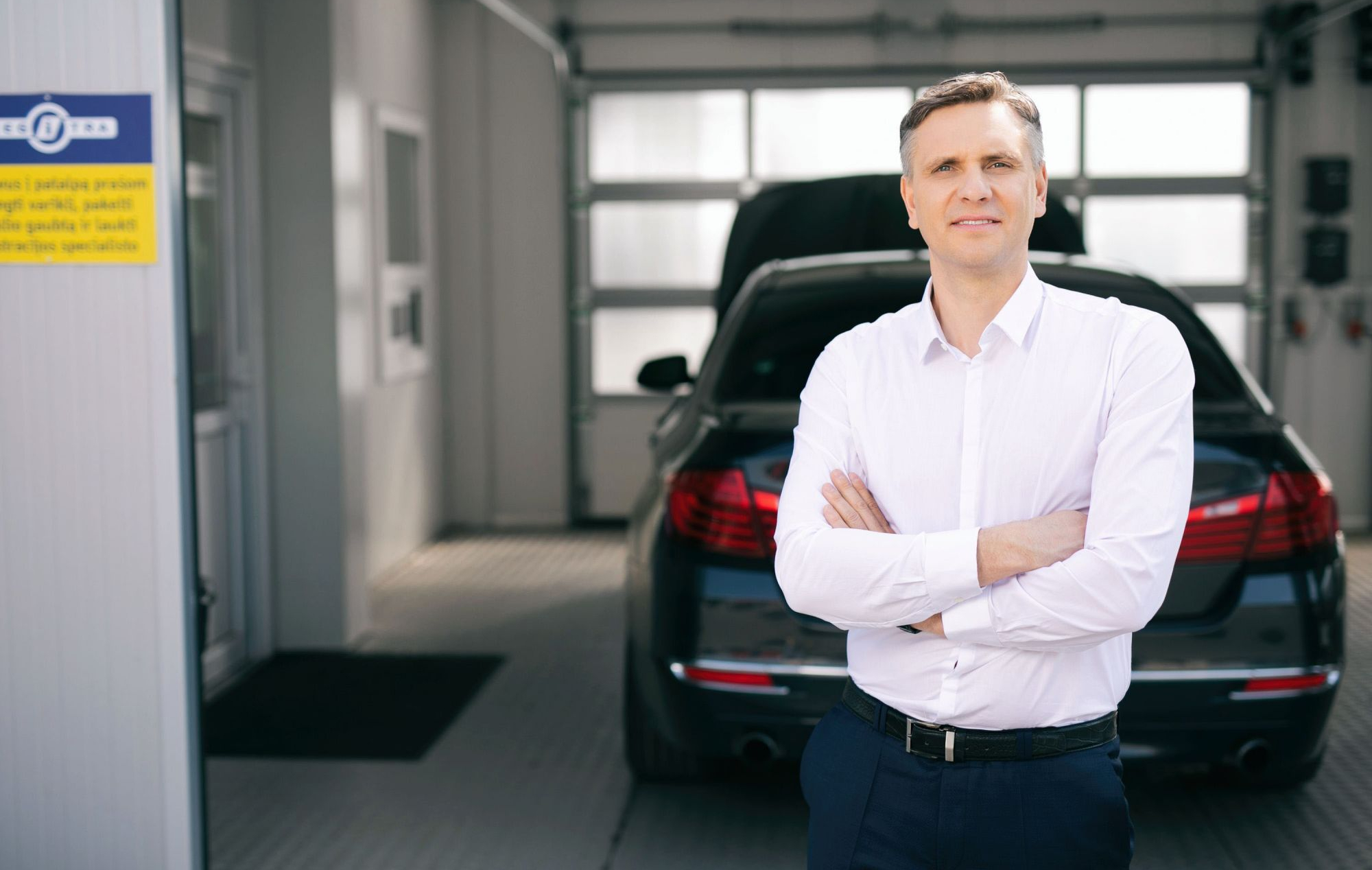 Asmuo su baltais marškiniais stovi sukryžiavęs rankas prie automobilio atidarytu kapotu.