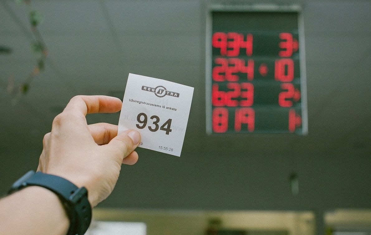 Paveikslėlyje matoma ranka, laikanti bilietėlį su numeriu „934“. Ant bilieto taip pat yra užrašai „REGITRA“ ir „Užsiregistravusiems iš anksto“. Fone matomas skaitmeninis ekranas, kuriame rodomi šie numeriai: 224 – 0 823 – 2 819 – 1