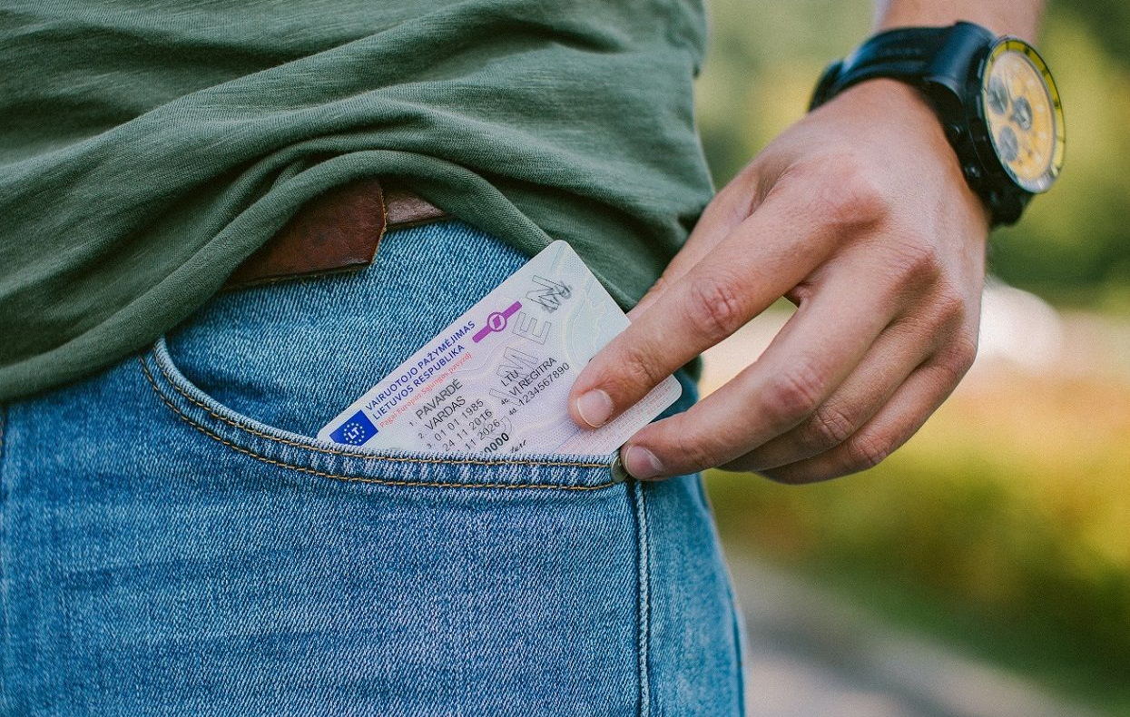 Asmuo su žaliais marškiniais ir mėlynais džinsais laiko vairuotojo pažymėjimą dalinai įkištą į priekinę kišenę. Matomas rudas diržas ir laikrodis ant kairės rankos.