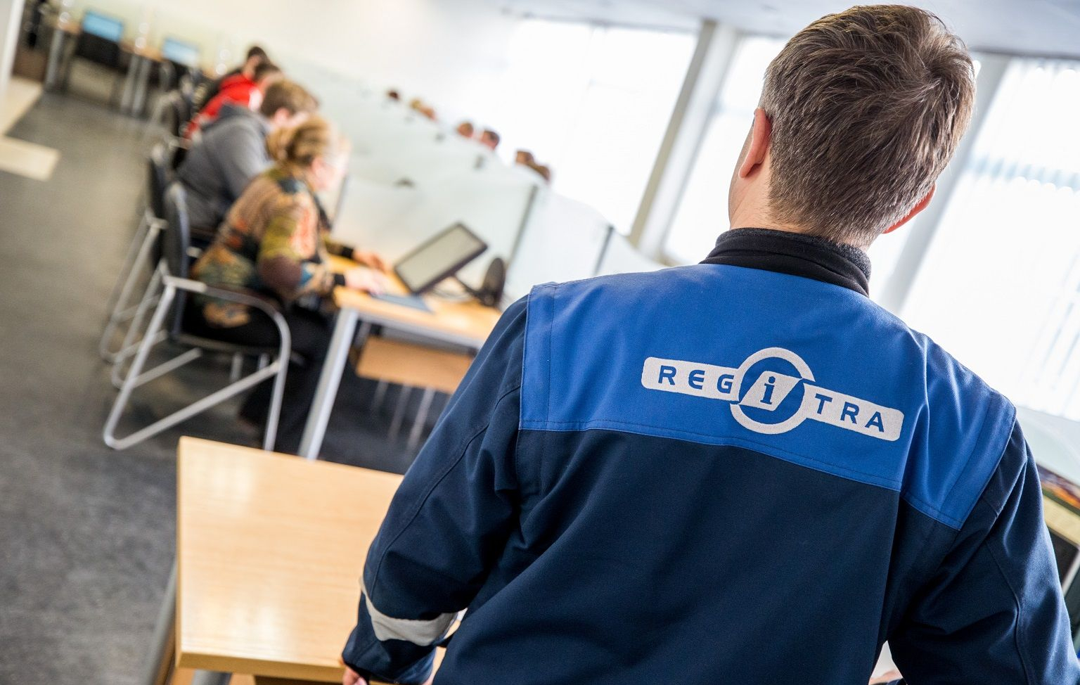 Darbuotojas su mėlyna striuke, ant kurios nugaros užrašyta „REGITRA“, stovi klasės aplinkoje. Fone keli žmonės laiko egzaminą su kompiuteriais prie stalų, patalpa apšviesta natūralia šviesa pro didelius langus.