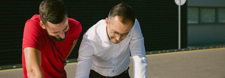 Du asmenys lauke pasilenkę ir žiūri į objektą už kadro. Vienas vilki raudonus marškinėlius, kitas – baltus.