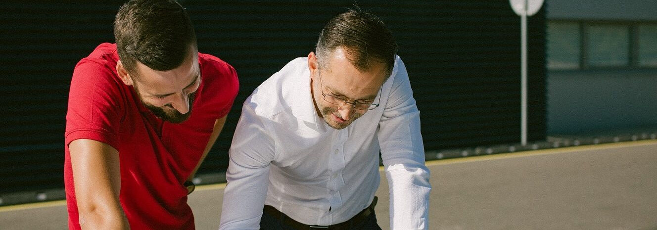 Du asmenys lauke pasilenkę ir žiūri į objektą už kadro. Vienas vilki raudonus marškinėlius, kitas – baltus.