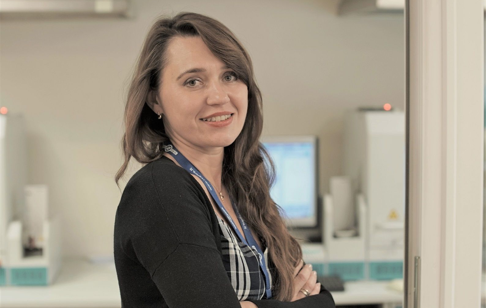 Darbuotoja su ilgais banguotais plaukais, vilkintis languotus marškinius ir juodą megztinį, stovi laboratorijos ar biuro aplinkoje. Fone matoma laboratorinė įranga ir kompiuterio monitorius.