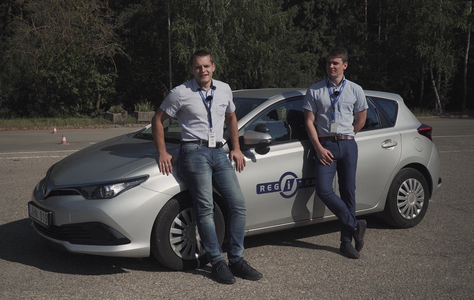 Regitros darbuotojai su šviesiai mėlynais marškiniais ir tamsiomis kelnėmis stovi prie sidabrinio automobilio su „REGITROS“ logotipu. Fone matomi medžiai ir atvira erdvė, aikštelė.