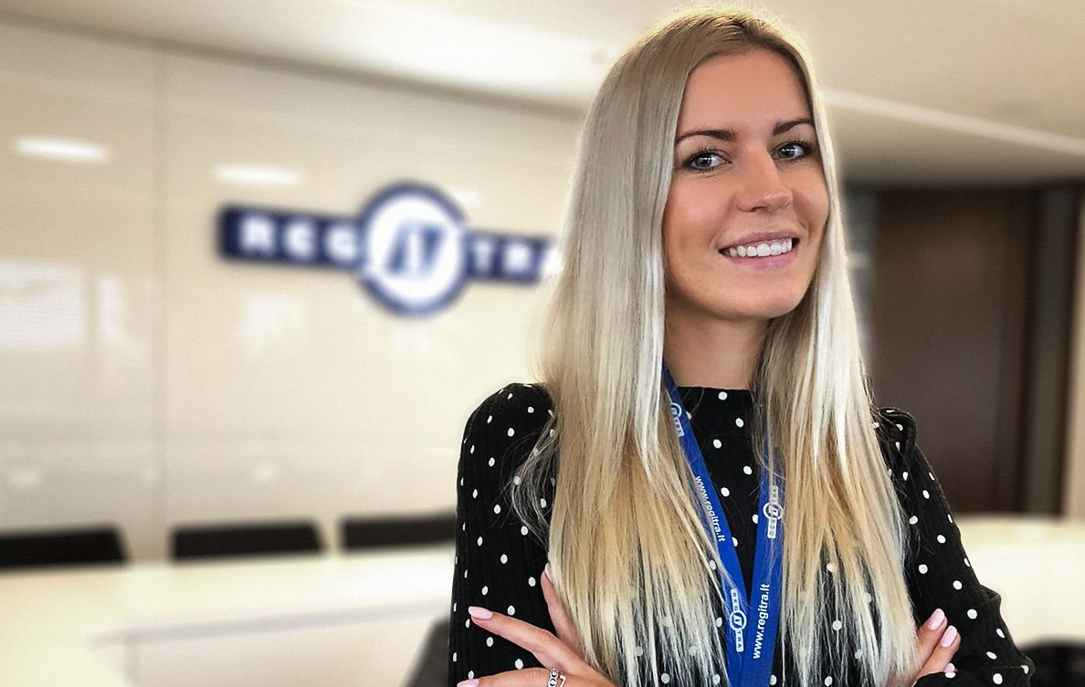 Moteris su ilgais šviesiais plaukais stovi biuro aplinkoje prie sienos su „Regitros“ logotipu. Vilki juodą suknelę su baltais taškeliais ir mėlyną kortelę ant kaklo