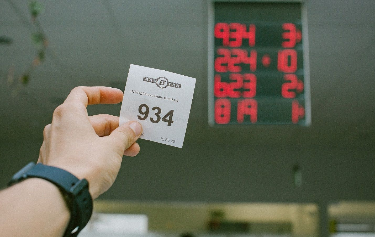 Ranka laiko bilietėlį su numeriu 934. Fone matomas elektroninis ekranas, rodantis numerių ir langelių atitikmenis: 934 – 3 langelis, 224 – 10, 623 – 2, 819 – 1. Bilietėlyje yra tekstas kita kalba ir laiko žyma „15:55:28“.