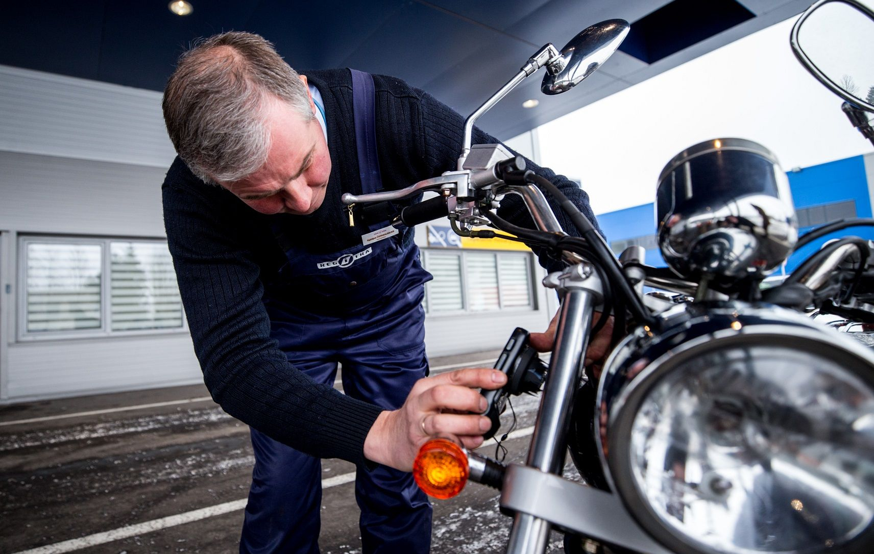 Asmuo su tamsiu megztiniu ir mėlynais darbo rūbais dirba prie motociklo priekinės dalies, šalia žibinto. Veidas užbluruotas privatumo sumetimais.