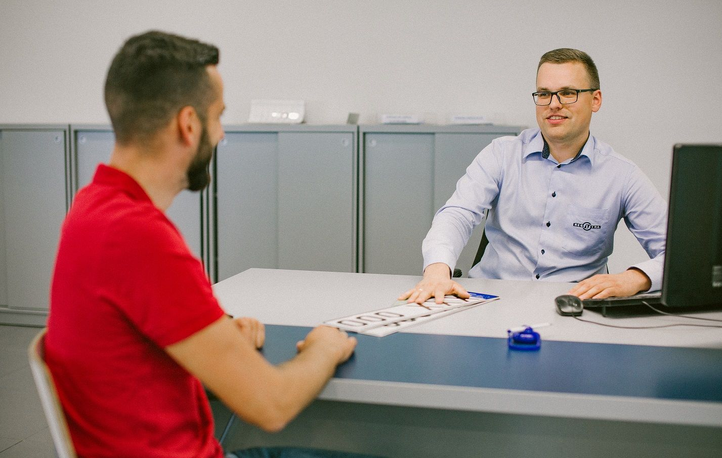 Du asmenys sėdi priešais vienas kitą biuro aplinkoje. Vienas vilki raudonus marškinėlius, kitas – šviesiai mėlynus marškinius ir akinius. Ant stalo yra kompiuterio monitorius, o asmuo su mėlynais marškiniais perduoda valstybinius numerius kitam.