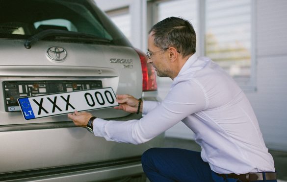 Asmuo su baltais marškiniais ir mėlynomis kelnėmis tvirtina arba nuima numerio lentelę nuo sidabrinio „Toyota“ automobilio. Numeris – XXX 000, su ES simboliu ir „LT“ žyma.