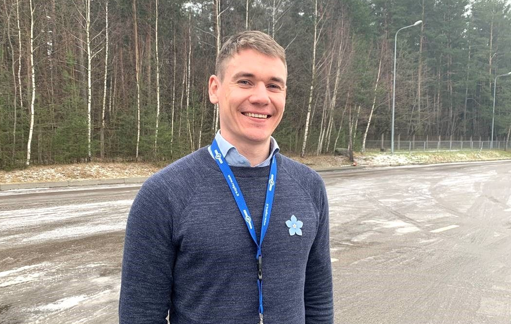 Asmuo su tamsiu megztiniu, šviesiai mėlynais marškiniais ir mėlynu pakabuku stovi lauke, fone – miškas ir sniegas