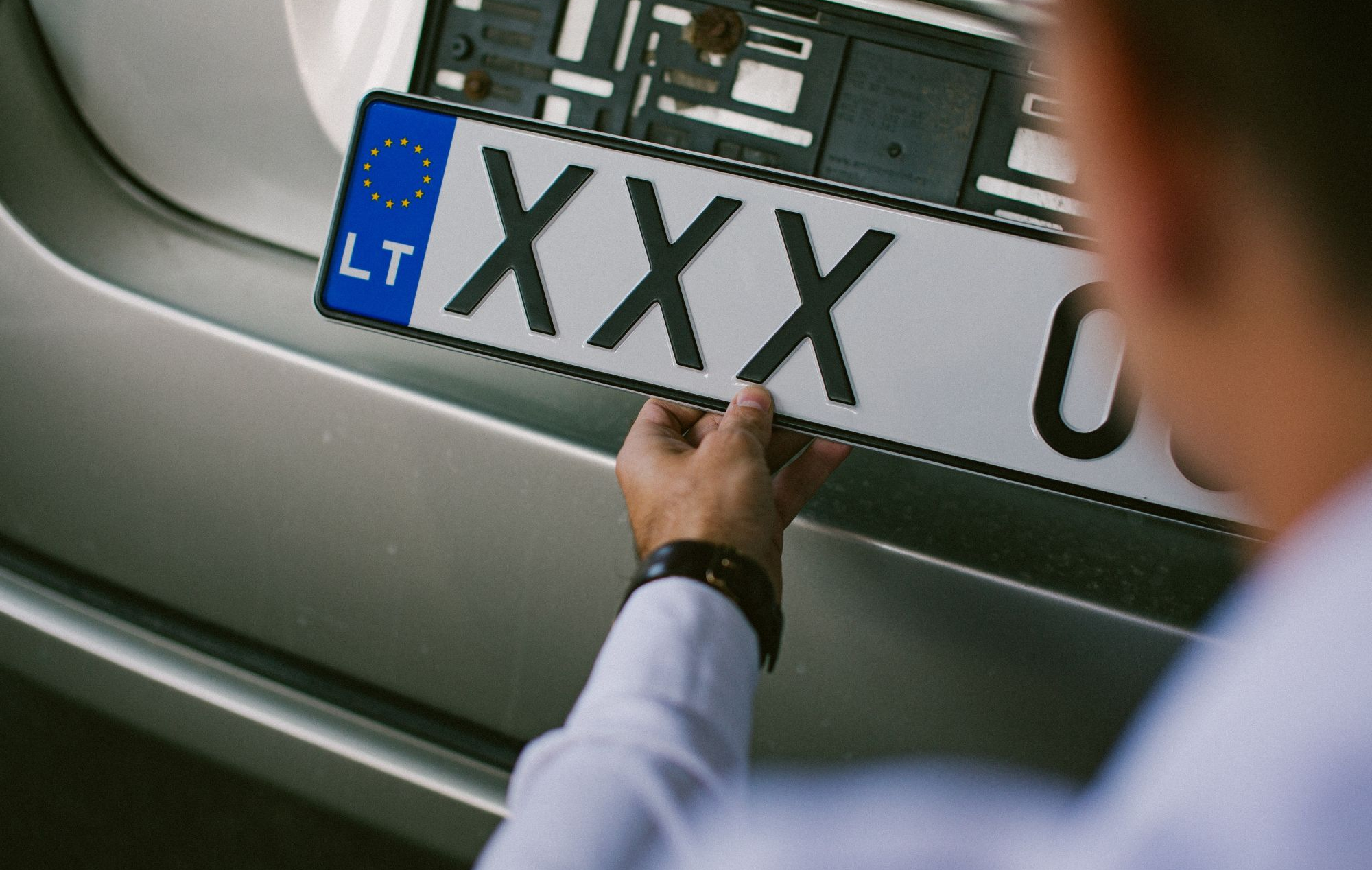 Asmuo laiko automobilio numerio ženklą su užrašu „XXX“ ir ES simboliu bei Lietuvos šalies kodu „LT“