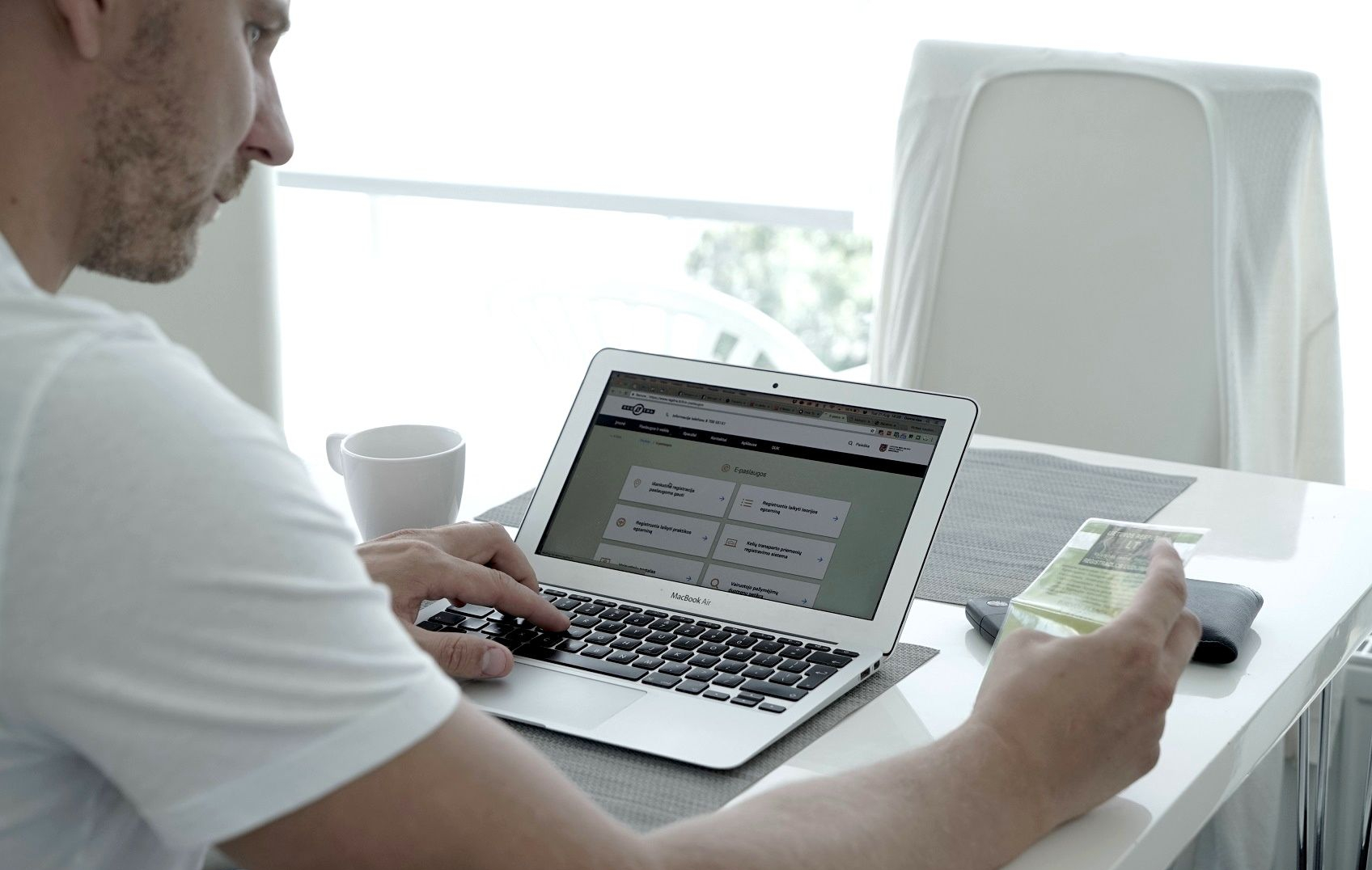 Asmuo sėdi prie stalo, viena ranka laiko išmanųjį telefoną, kita rašo „MacBook“ kompiuteriu, šalia – balta kavos puodelis