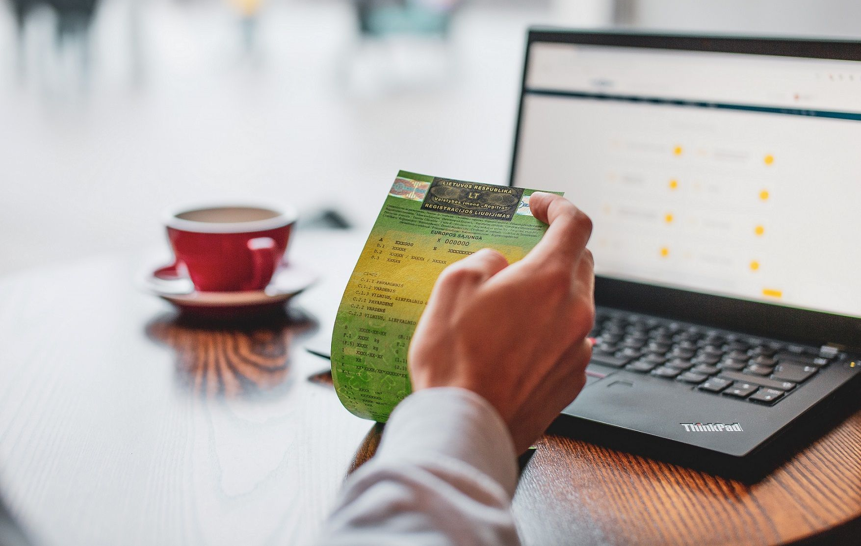 Nuotraukoje matomas žmogus, laikantis registracijos dokumentą priešais nešiojamąjį kompiuterį, kuris stovi ant medinio stalo.