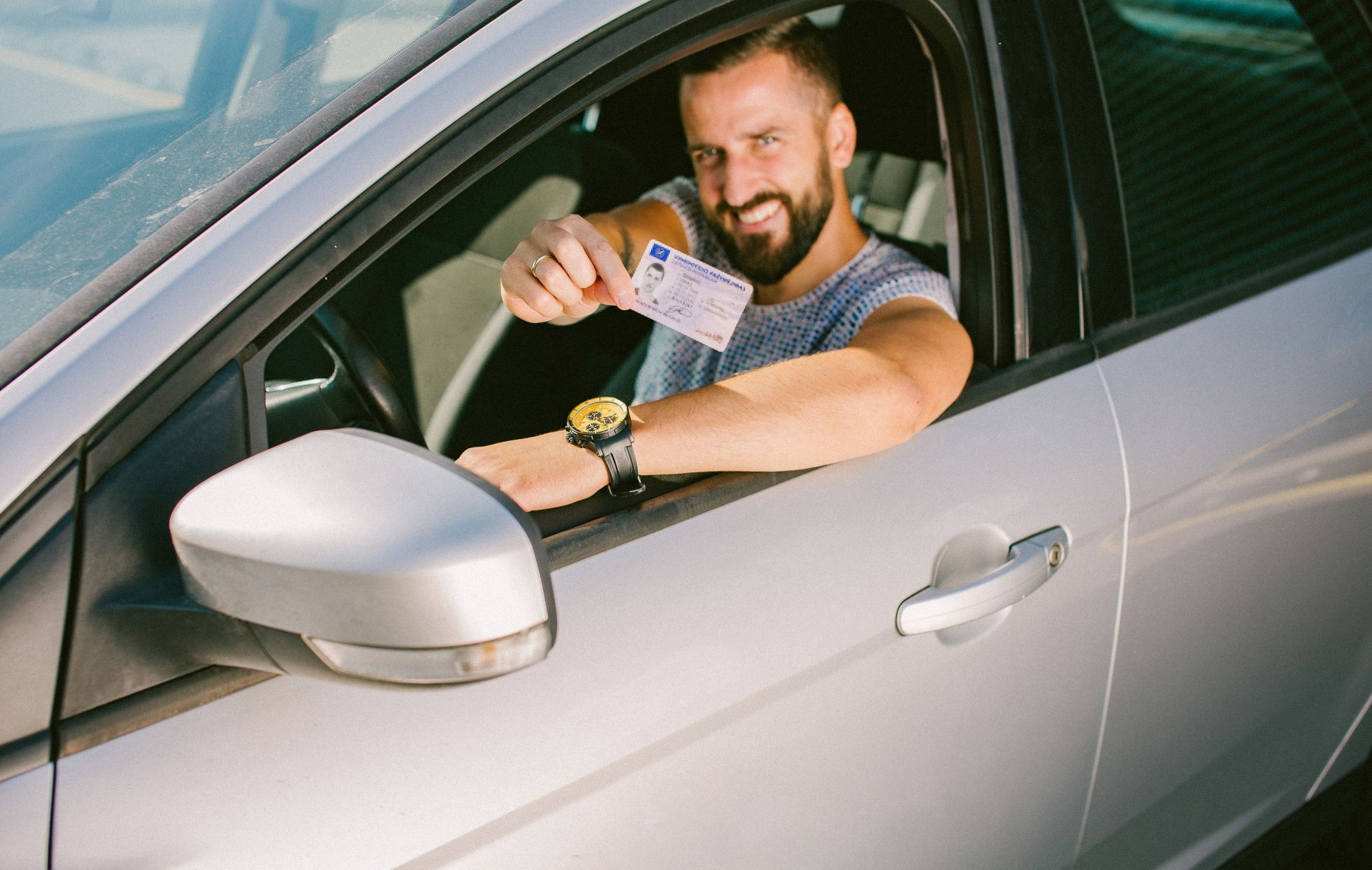 Asmuo sėdi sidabrinio automobilio vairuotojo vietoje ir pro atvirą langą laiko vairuotojo pažymėjimą.