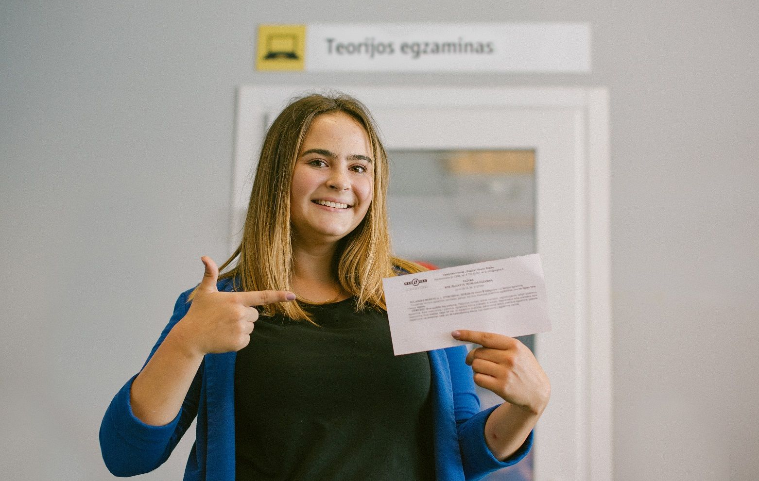 Šioje nuotraukoje matyti žmogus, stovintis priešais teorijos egzamino klasę ir laikantis lapelį ant kurio parašyta išlaikytas teorijos egzaminas.