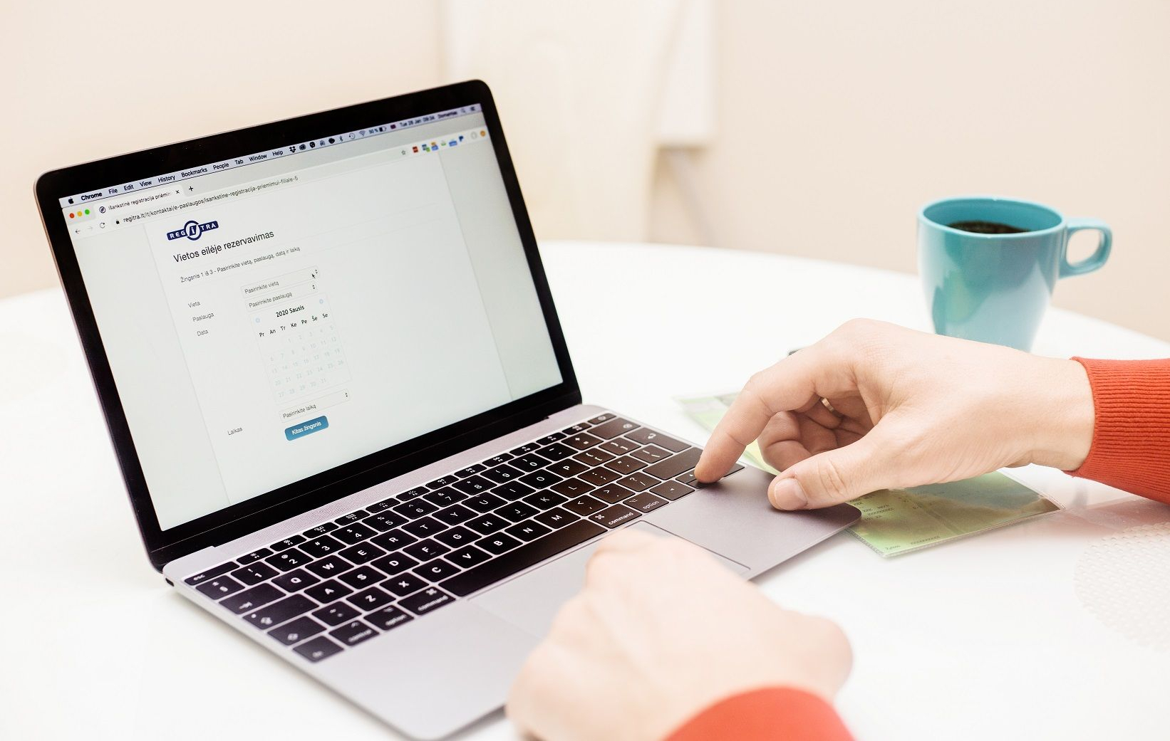 Nuotraukoje matomas žmogus, naudojantis nešiojamąjį kompiuterį ant balto stalo. Ekrane atidaryta internetinė forma su antrašte „Vieta online rezervavimas“, kurioje yra laukeliai pasirinkti „Lytis“, „Amžius“ ir „Data“, su išskleidžiamais meniu ir kalendoriumi. Taip pat matomas mėlynas mygtukas „Pasirinkti vietą“. Dešinėje stalo pusėje padėtas mėlynas puodelis.