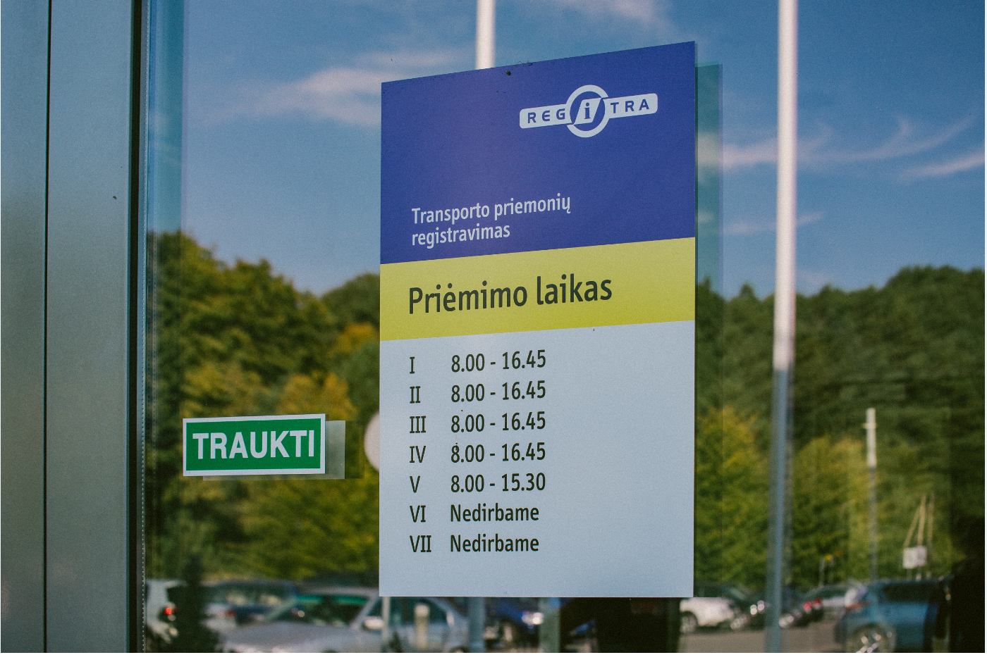 Šioje nuotraukoje matomas informacinis stendas ant stiklinės durų su „REGITRA“ logotipu viršuje. Po juo pateikta informacija apie transporto priemonių registravimo darbo laiką: I–IV: 8.00 – 16.45 V: 8.00 – 15.30 VI–VII: nedirbama