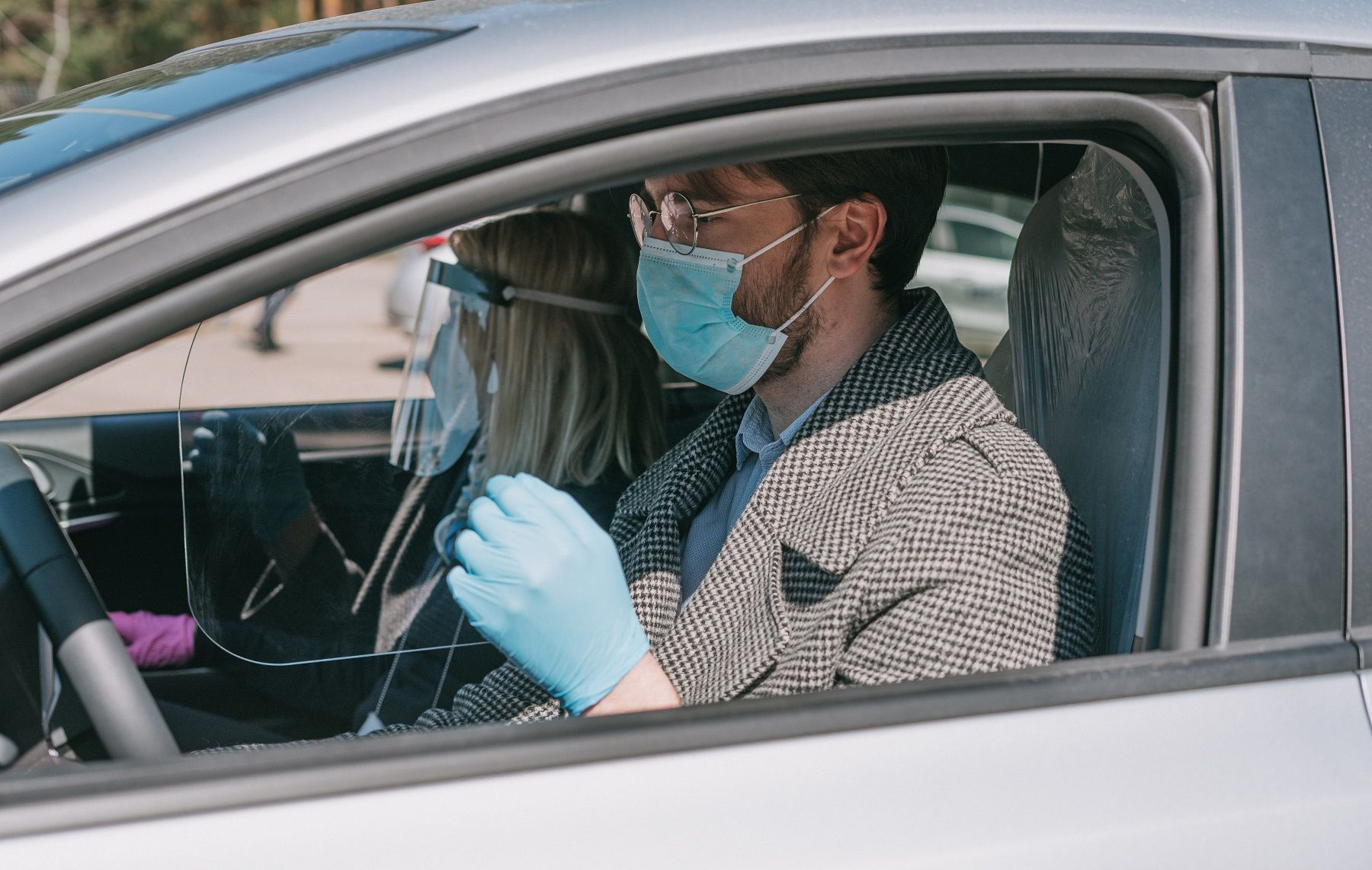 Matomi du žmonės automobilio priekyje, egzaminuotojas ir egzaminuojamasis. Vairuotojas dėvi veido kaukę, o keleivis – kaukę, pirštines ir languotą paltą.