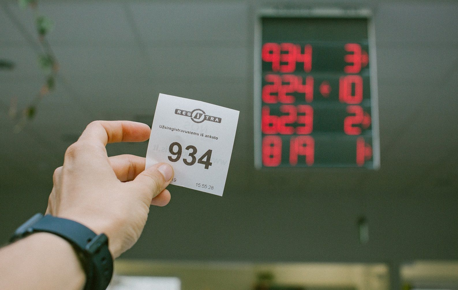 Ranka laiko bilietėlį su numeriu 934. Fone matomas elektroninis ekranas, rodantis numerių ir langelių atitikmenis: 934 – 3 langelis, 224 – 10, 623 – 2, 819 – 1. Bilietėlyje yra tekstas kita kalba ir laiko žyma „15:55:28“.