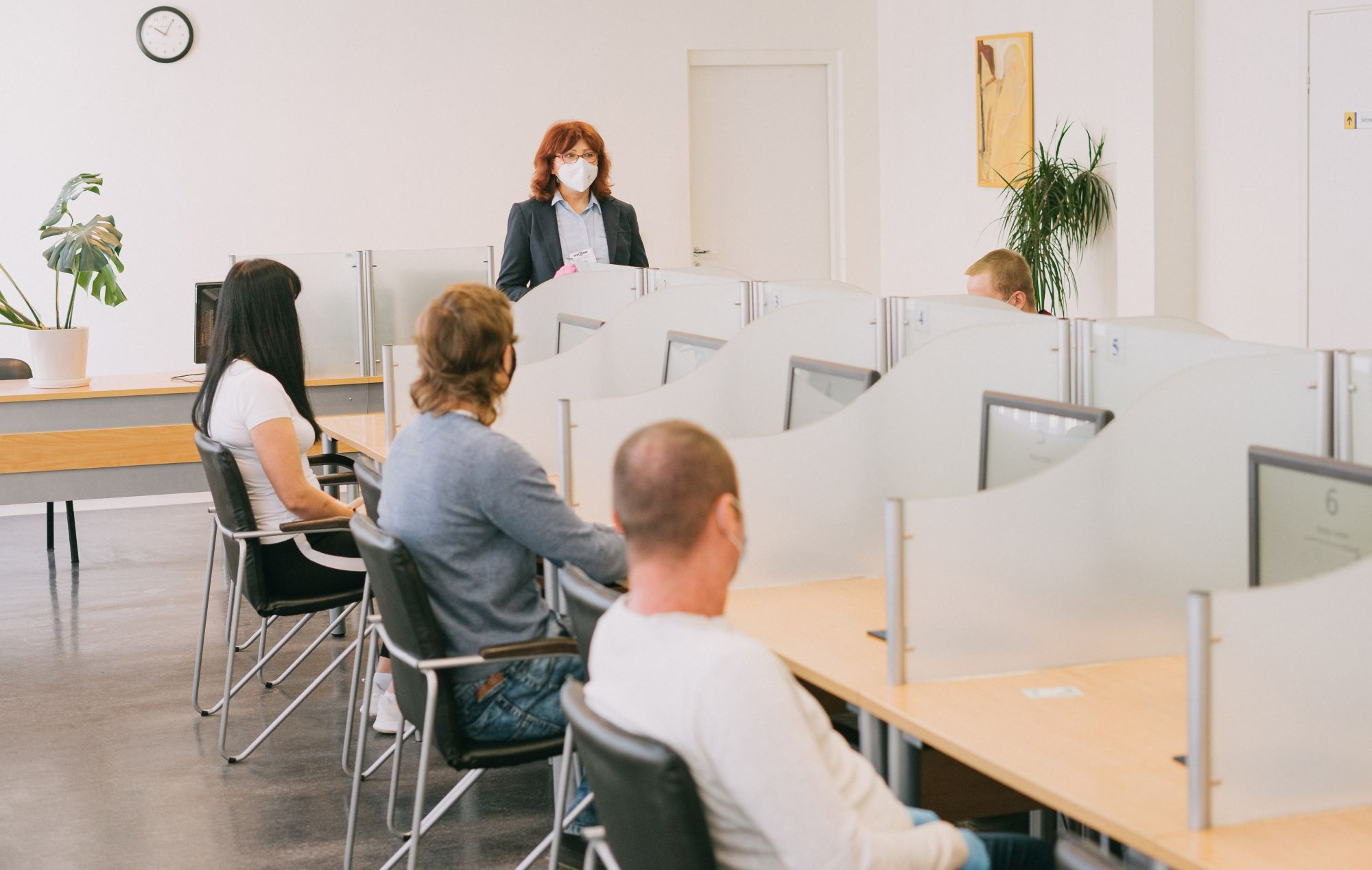 Nuotraukoje matoma teorijos egzamino patalpa, kurioje keli žmonės sėdi prie atskirų darbo vietų su kompiuteriais. Priekyje stovi darbuotojas. Darbo vietos atskirtos matinio stiklo pertvaromis. Ant sienos kabo laikrodis, o patalpoje taip pat yra augalų.
