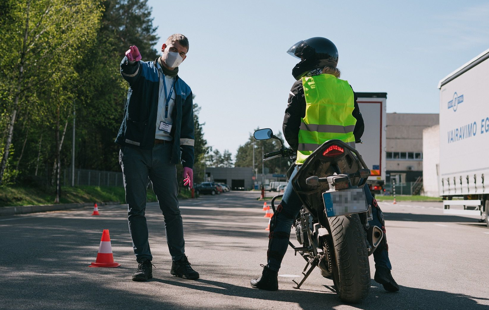 Nuotraukoje matomas žmogus su šalmu ir ryškiaspalve liemene, sėdintis ant motociklo. Šalia stovi egzaminuotojas, dėvintis veido kaukę ir pirštines, kuris rodo į kažką aplinkoje.. Fone matyti pastatai ir transporto priemonės.