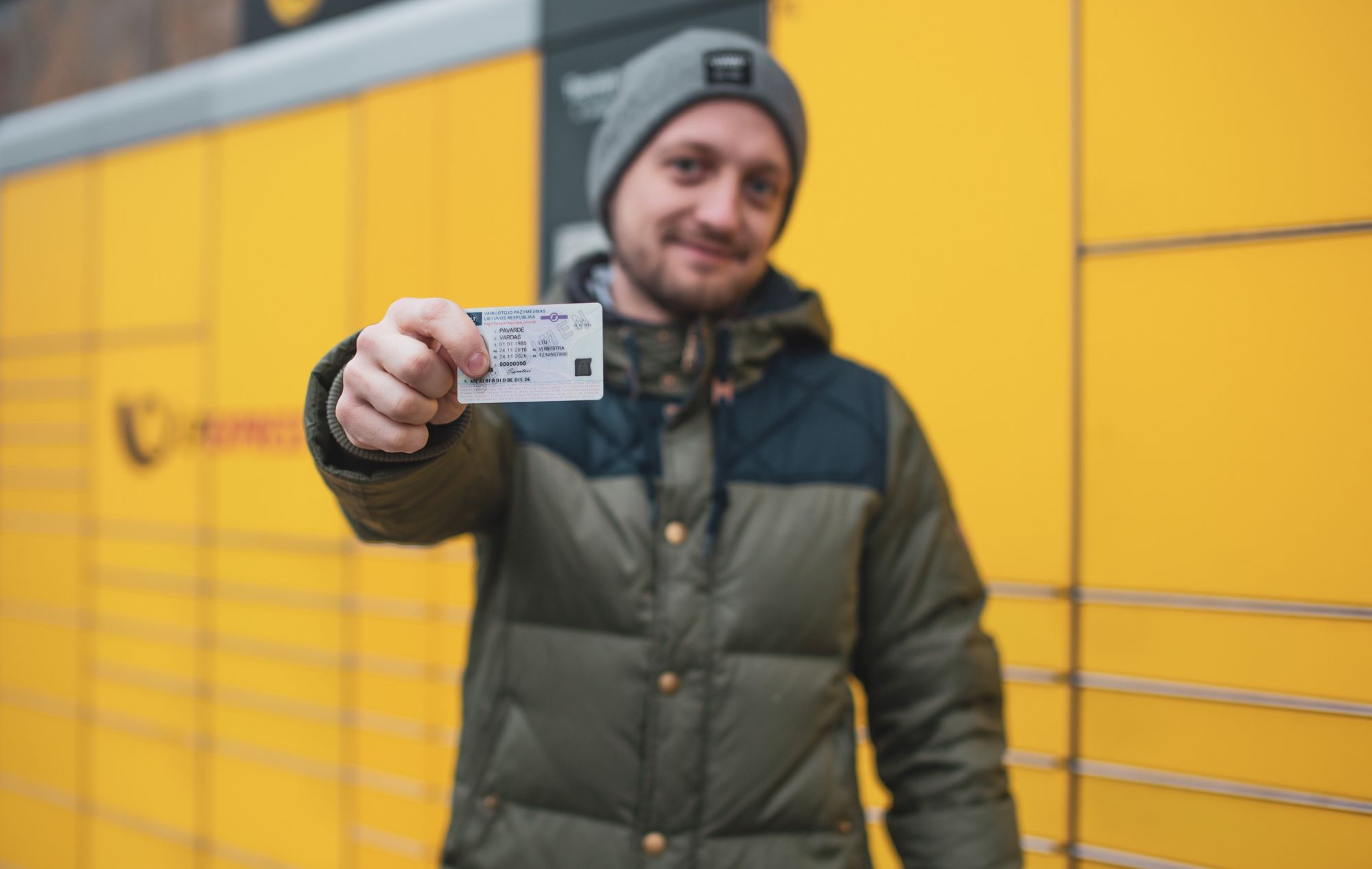 Matomas žmogus su žaliai juodu švarku ir pilka kepure, stovintis prieš geltoną foną su horizontaliomis linijomis. Veidas yra sulietas, o žmogus laiko asmens tapatybės kortelę, nukreiptą į kamerą.