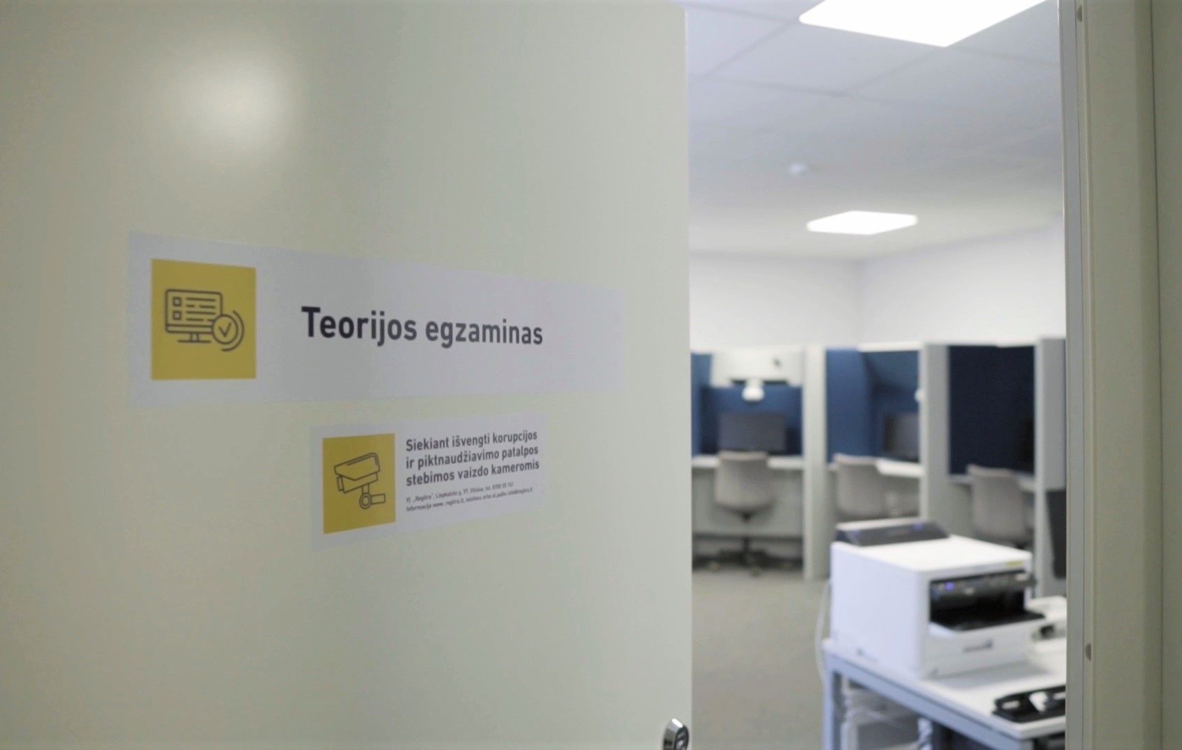 Nuotraukoje matomos durys, vedančios į patalpą su keliais kompiuteriais. Ant durų yra du ženklai: „Teorijos egzaminas“ – su dokumento ir varnelės ikona. „Siekiant išvengti korupcijos ir piktnaudžiavimo patalpos stebimos vaizdo kameromis“ – su stebėjimo kameros ikona.