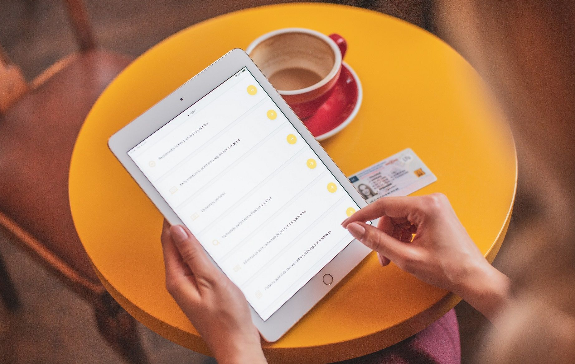 Asmuo prie geltono stalo naudoja planšetę, šalia – raudonas puodelis su lėkštute ir asmens tapatybės kortelė.