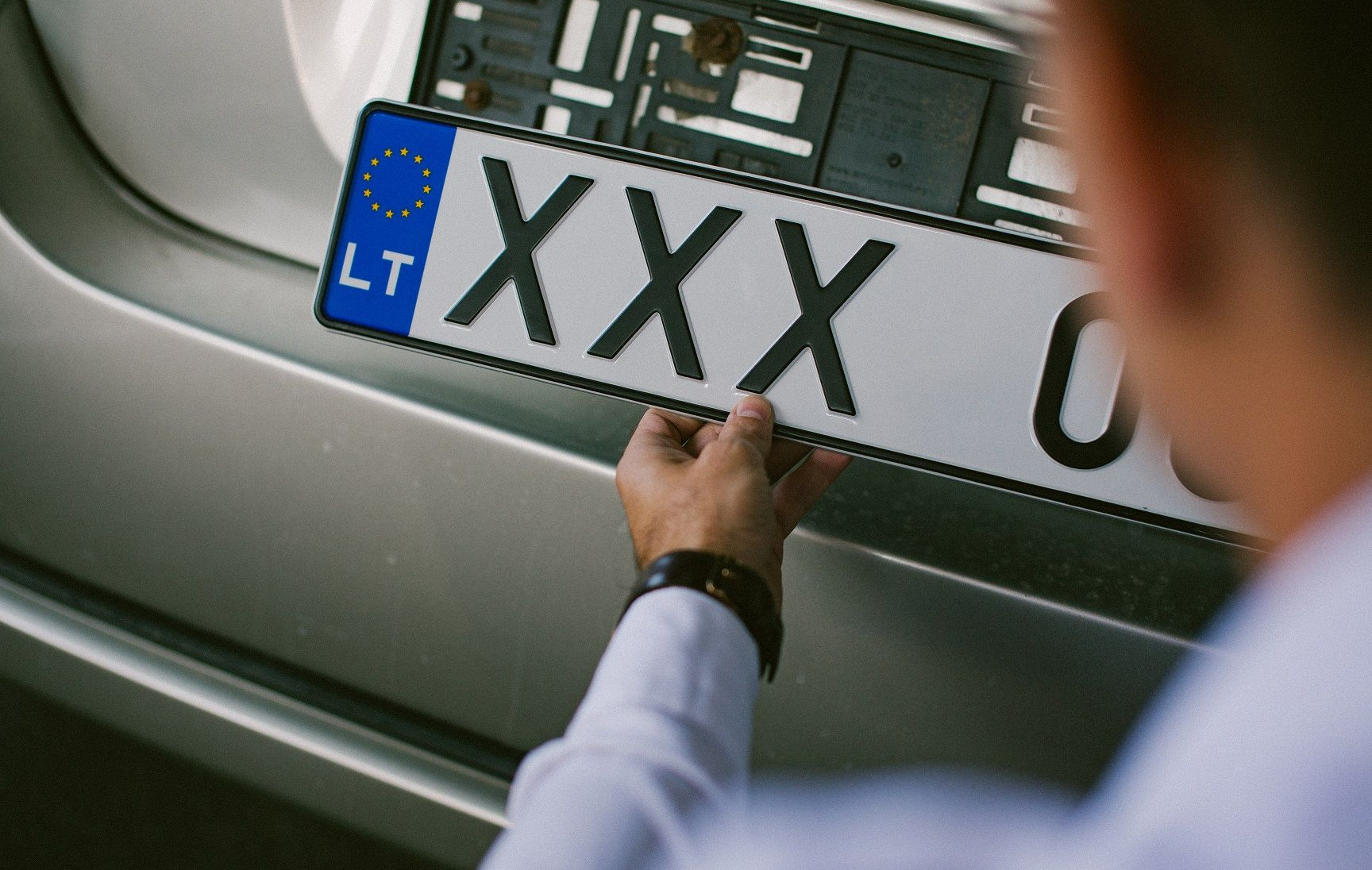 Asmuo laiko lietuvišką automobilio numerio lentelę su užrašu „XXX“, šalia matomas automobilio galas.
