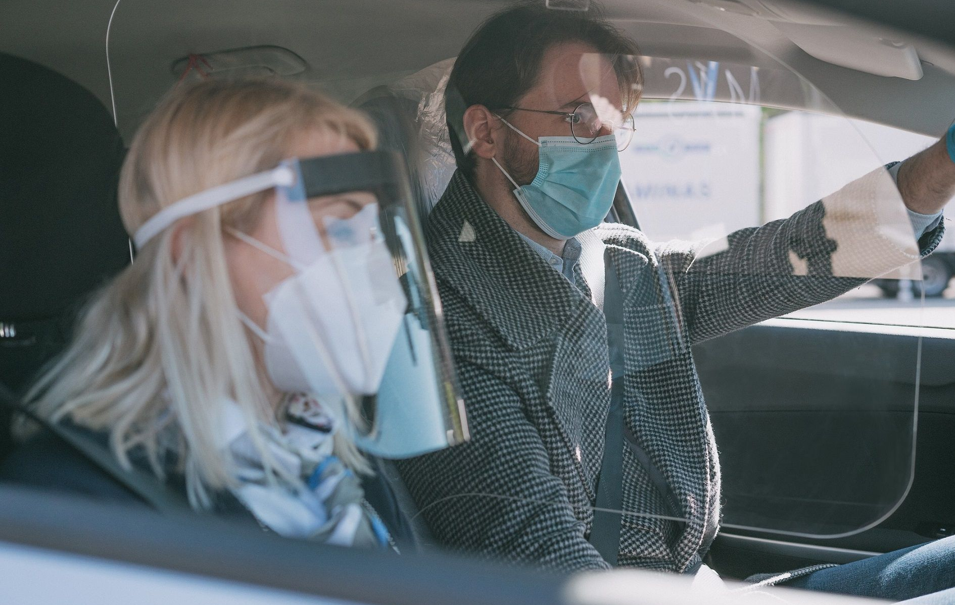 Du žmonės automobilyje – vairuotojas su apsaugine kauke ir keleivis, abiejų veidai užbluruoti.